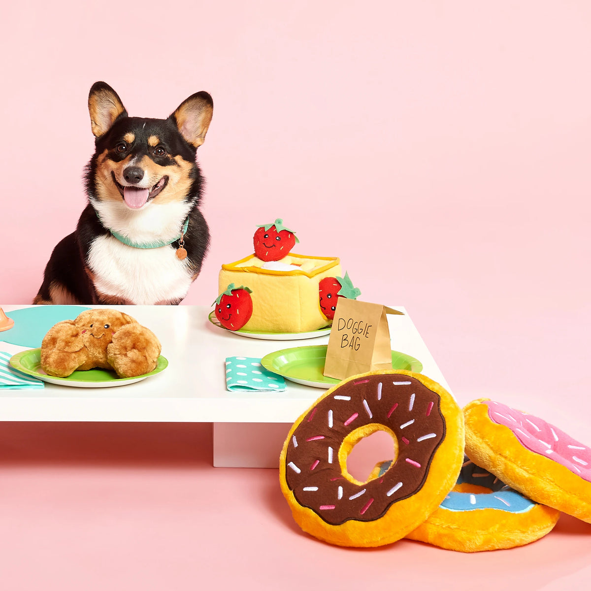 Corgi seated at a table laid with Zippy Paws plush food dog toys including Donutz Jumbo