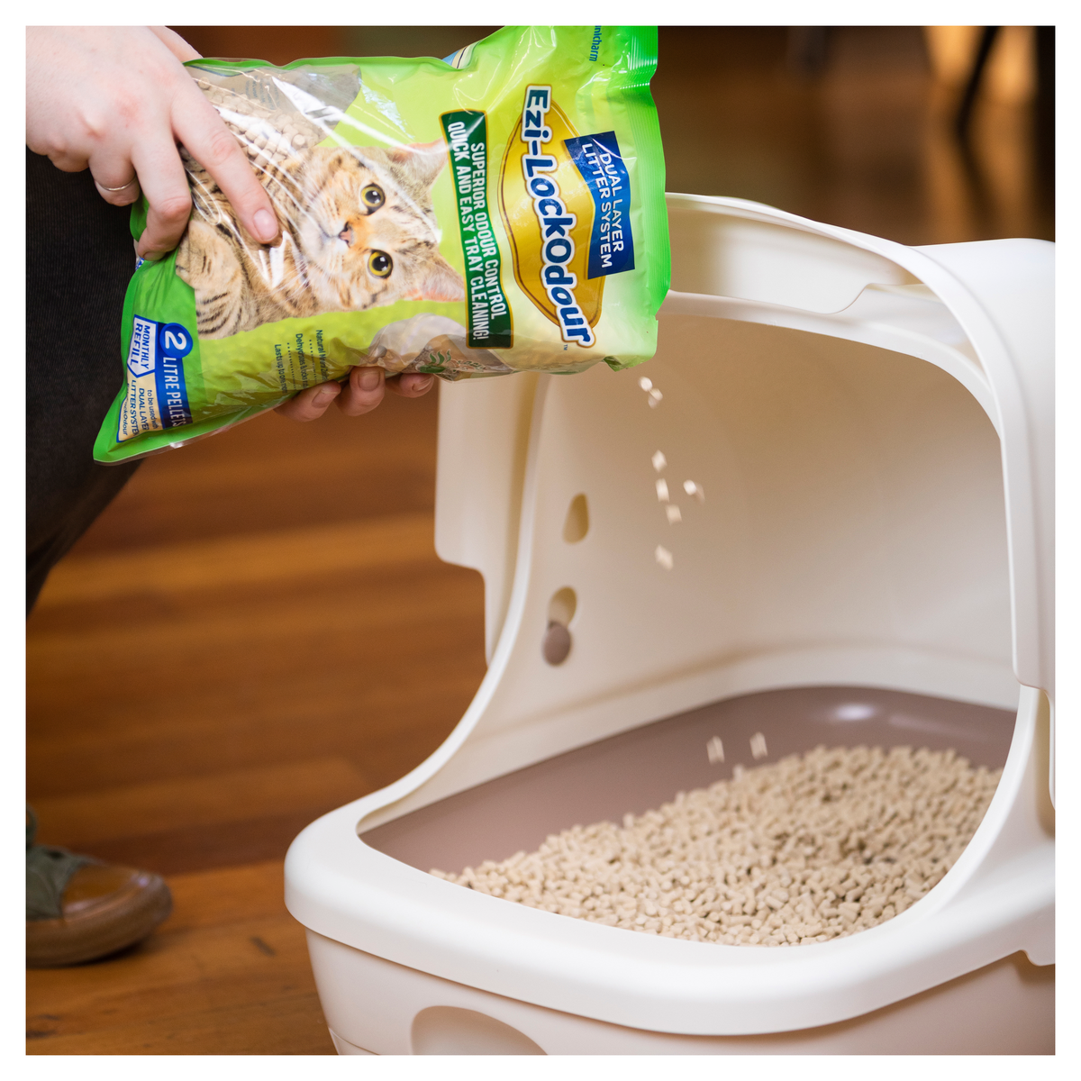 Hands pouring zeolite pellets into the open tray of an Ezi-LockOdour litter box