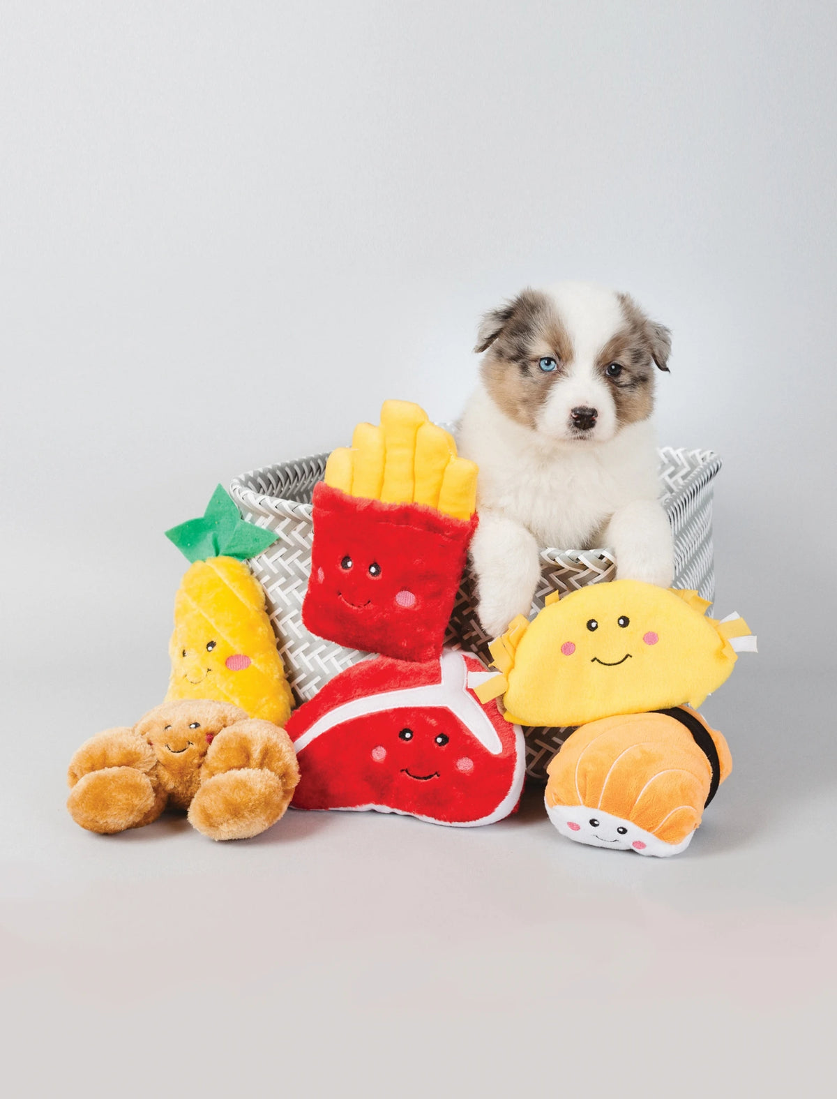 Puppy in a basket surrounded by Zippy Paws NomNomz plush food dog toys
