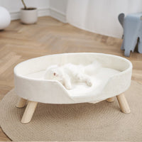 A white cat lying in the Petsbelle Snowmelt cat bed, a cream curved bed raised on timber legs