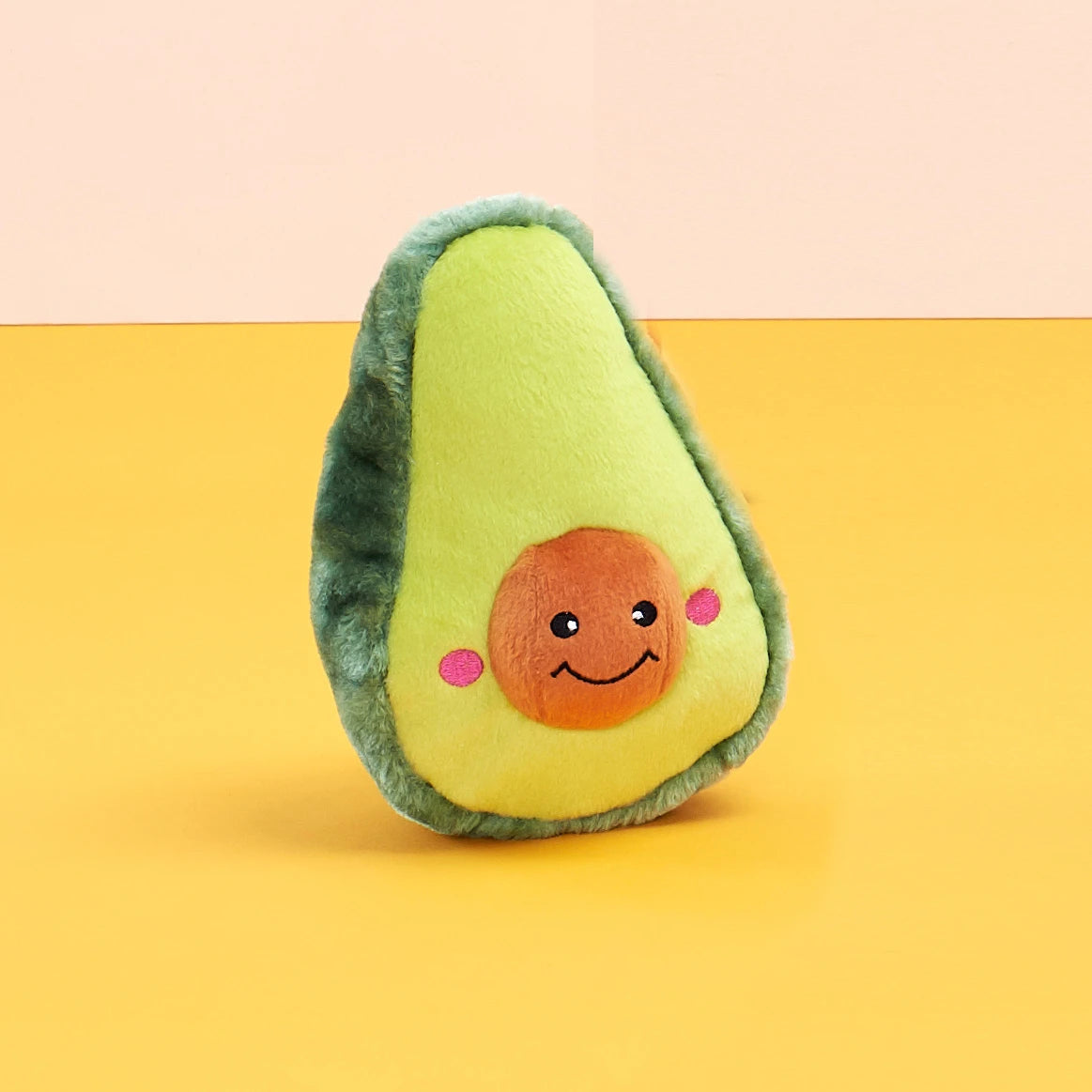 Zippy Paws NomNomz plush avocado dog toy styled on a yellow and peach backdrop